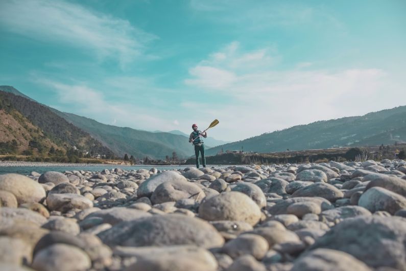 Bhutan Landscape - man holding oar standing on stones near body of water during daytime