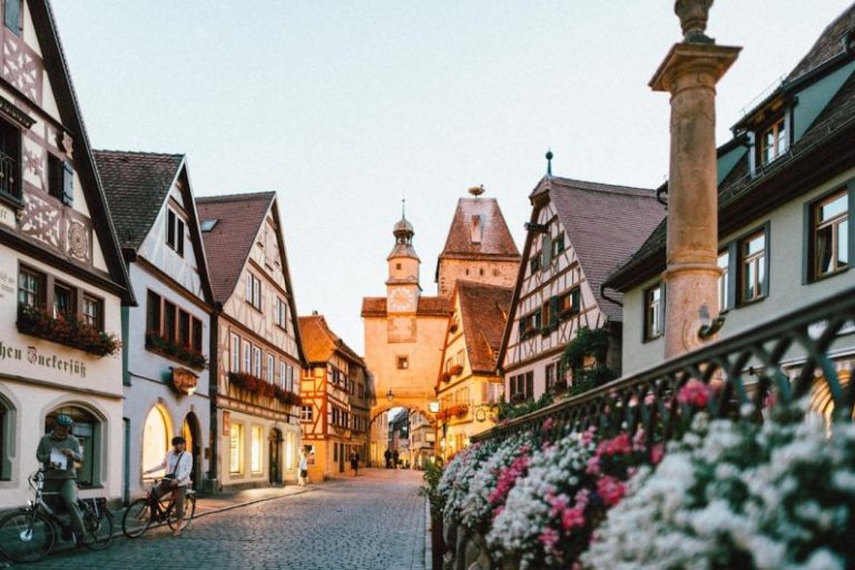Europe Travel - white and pink petaled flowers on metal fence near concrete houses and tower at daytime