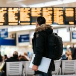 Travel Insurance - man standing inside airport looking at LED flight schedule bulletin board